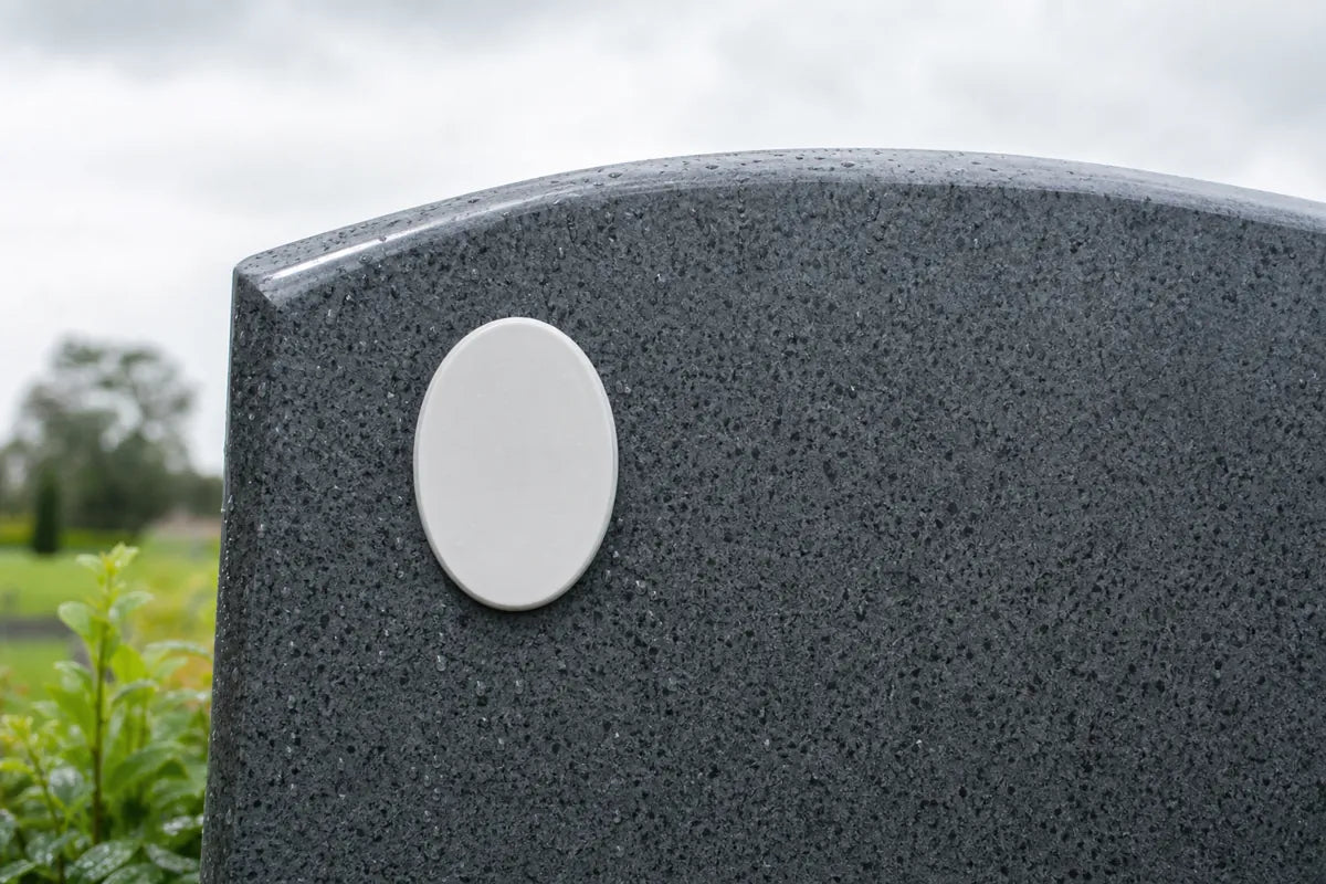 Illustration of a blank white oval plaque on a rain-dotted dark granite headstone under an overcast sky
